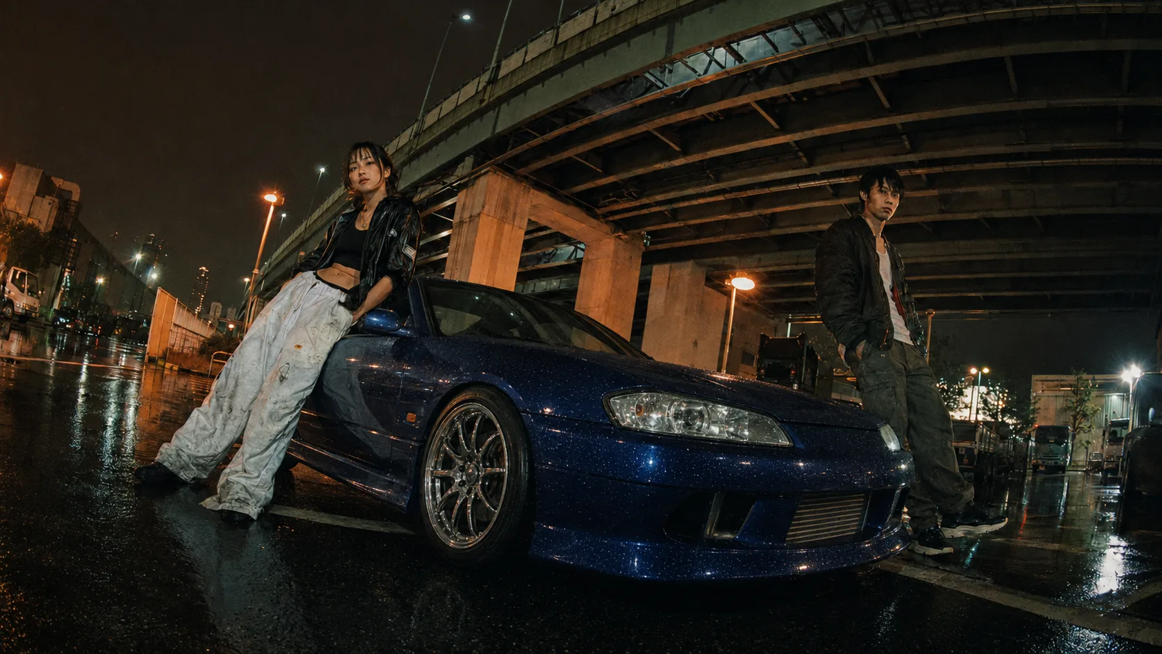 Two people in oversized jackets stand either side of a low blue coupe on wet ground beneath a curving elevated expressway at night.